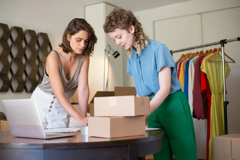 Two women in a small clothing shop packing cardboard boxes at a round table with a laptop, with a clothes rail behind them