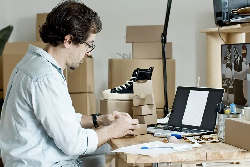 Seller at a desk packing a small parcel beside a laptop, with sneaker boxes stacked behind him