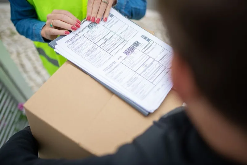 Courier in a high-visibility vest holding delivery paperwork on top of a cardboard parcel during a handover