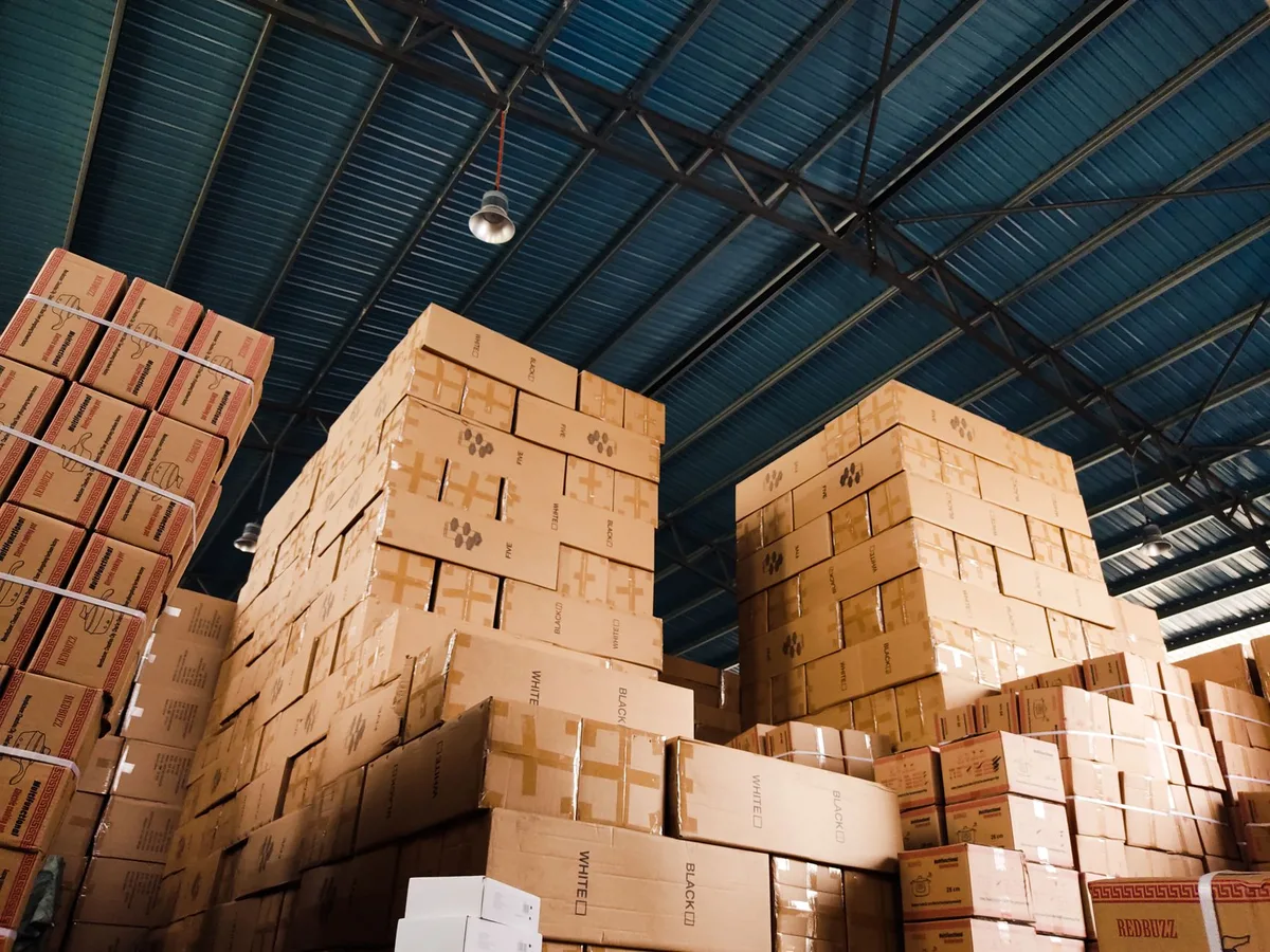 Tall stacks of cardboard boxes in a warehouse under a blue steel roof
