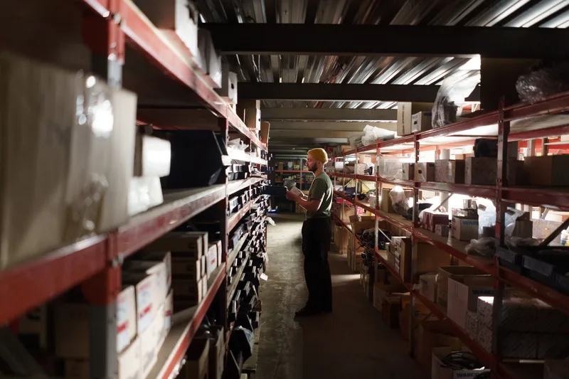 Worker in a dim warehouse aisle checking stock on red shelving racks