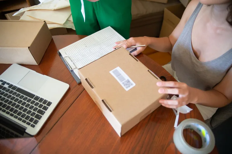 Seller packing a labelled cardboard order at a desk beside a laptop and a paper checklist