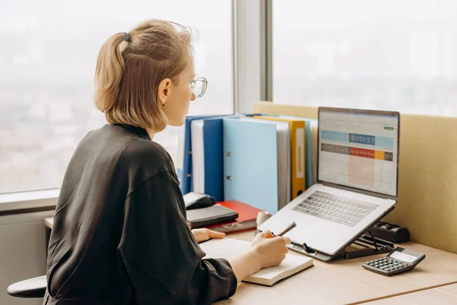 Woman at a desk making notes beside a laptop that shows a colourful spreadsheet
