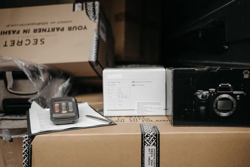 Handheld terminal and clipboard resting on cardboard parcels next to a camera box, ready for shipment