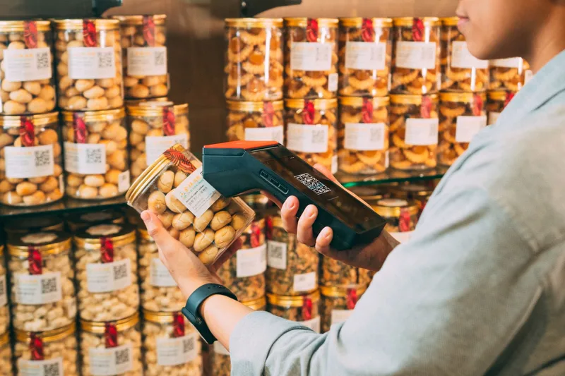 Shop worker scanning the QR label on a jar of snacks with a handheld scanner, with rows of labelled jars on the shelves behind