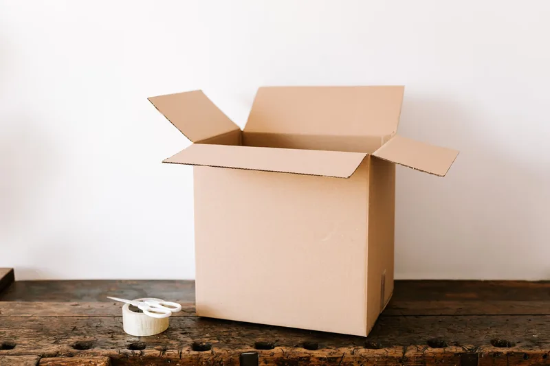 Open cardboard box on a wooden bench beside a roll of tape and scissors