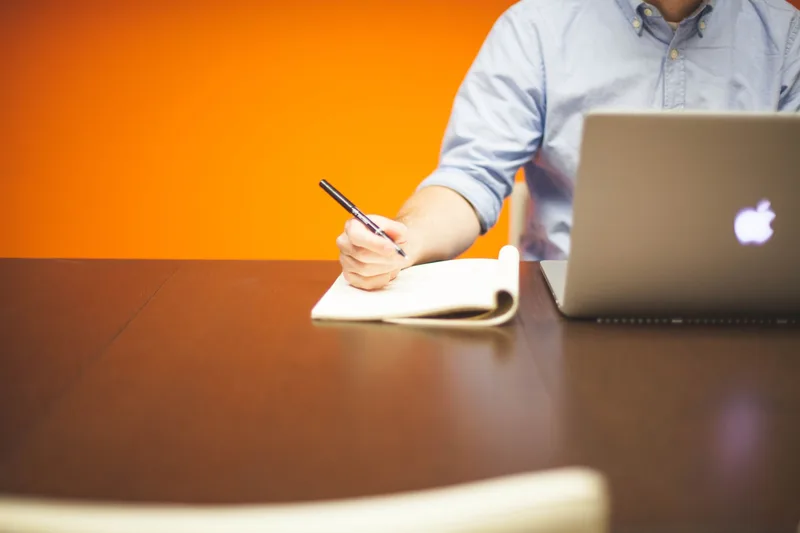 Person writing notes beside a laptop on a desk