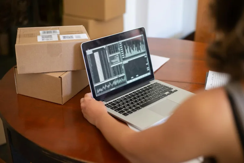 Small business owner reviewing ecommerce orders on laptop alongside shipping boxes — the operational context where WMS warehouse management software adds structure